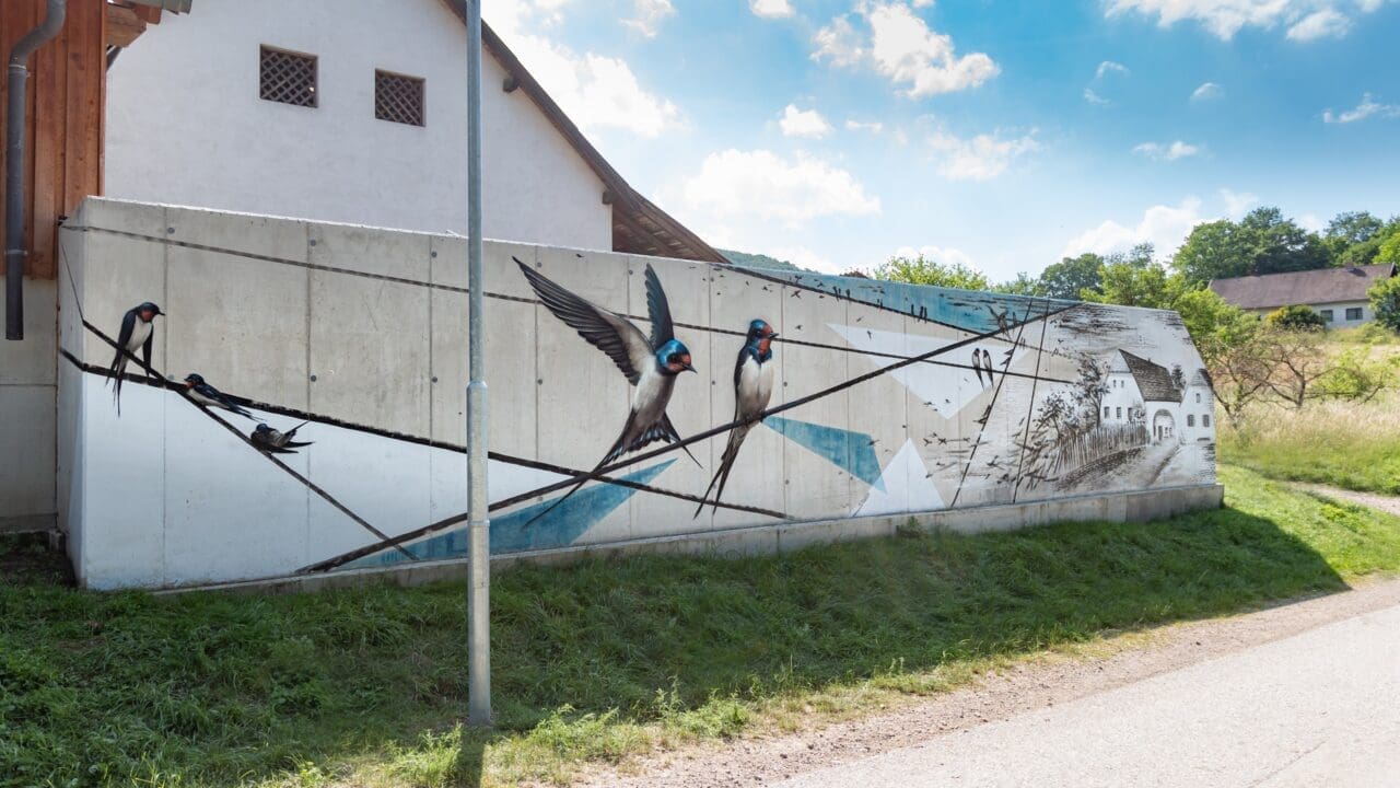 Wandkunstwerk auf Stahlbetonmauer mit Schwalben und Haus