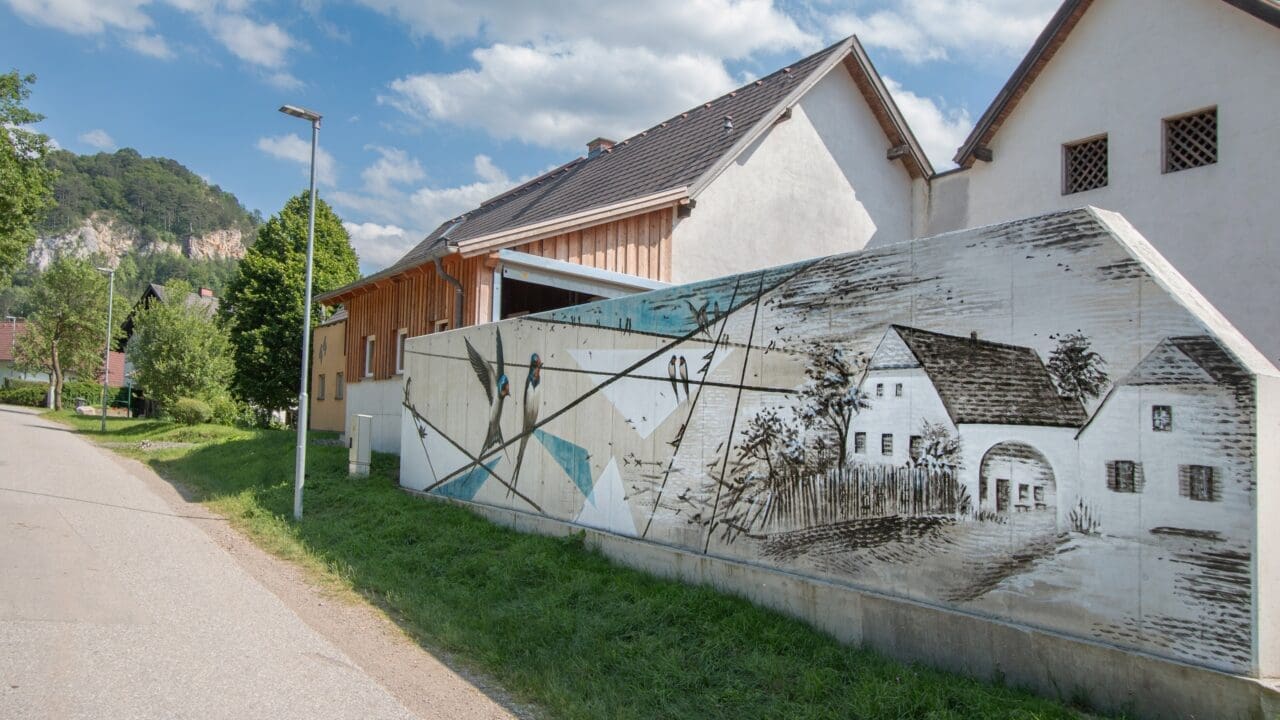 Wandkunstwerk auf Stahlbetonmauer mit Schwalben und Haus